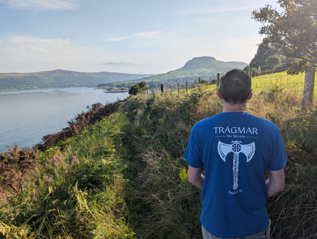 A man wearing a Tragmar Ale Works T-shirt stands facing a scenic view of fields and mountains along the water.