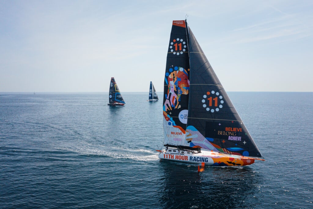 Three sailboats on the ocean. The one in front has black sails with a colorful design on them.