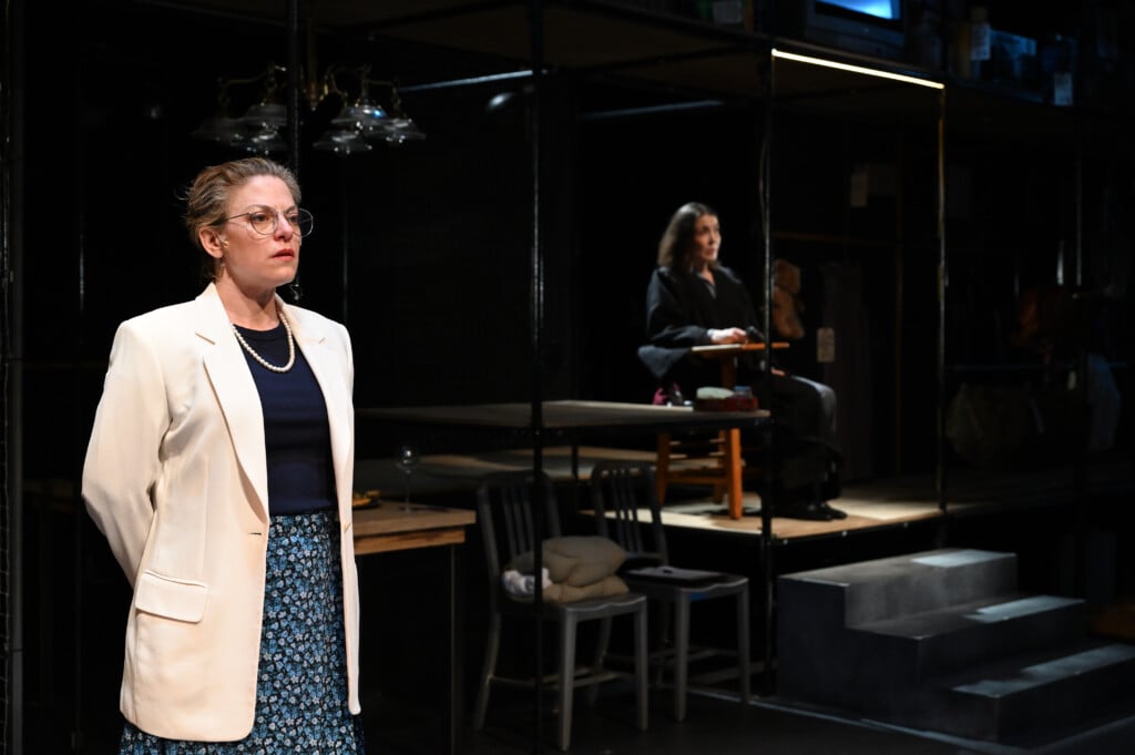 A woman stands in a white jacket in the foreground of a theater set while a woman in dark clothing sits in the background.