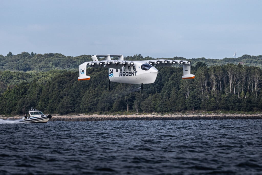 A white REGENT Seaglider flies over the water with trees visible on the shore in the background.