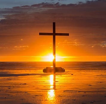 Cross On A Beach With The Sun Setting Down Behind It.